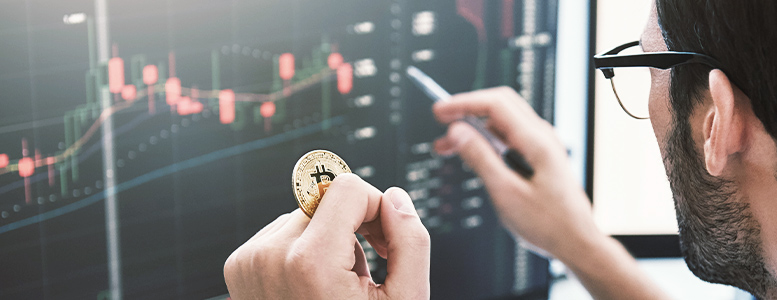 An investor holds a physical bitcoin token while staring at financial graphs on his computer monitors.