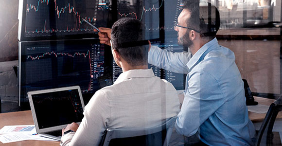 Two men study multiple computer screens with different types of financial graphs. 