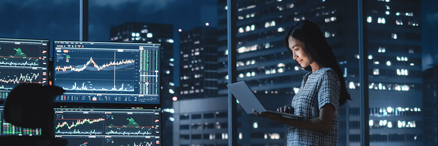 A person stands in an office at night looking at a laptop computer and surrounded by data charts on multiple monitors.