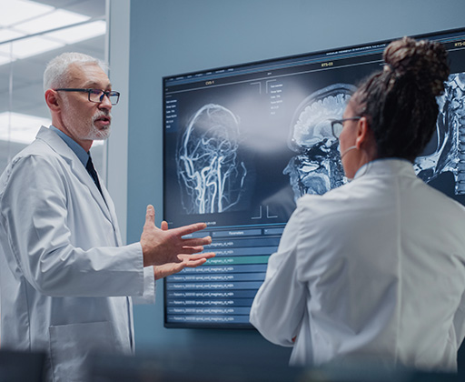 Two medical professionals in lab coats discussing x-ray charts on a wall lightboard.